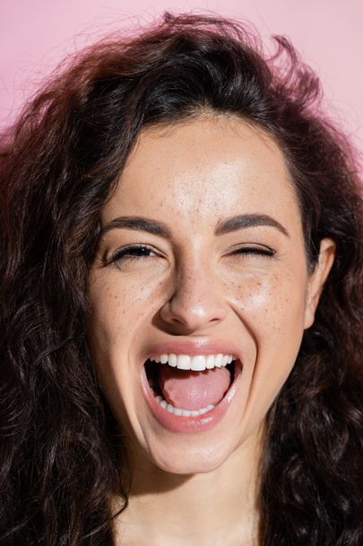 Portrait of excited and curly freckled woman looking at camera isolated on pink  - Фото, зображення