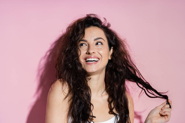 Positive brunette woman touching curly hair on pink background  - Фото, изображение