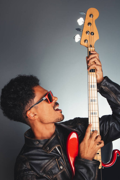 Handsome Black Musician Man With Leather Jacket Holding Bass. Portrait of a Man Wish Red Glasses Holding a Instrument Isolated. - Fotografie, Obrázek