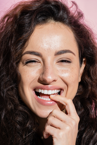 Positive curly woman looking at camera and holding finger near teeth isolated on pink  - Foto, immagini
