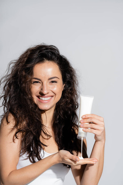 Smiling curly woman pouring body lotion on hand isolated on grey  - Foto, immagini