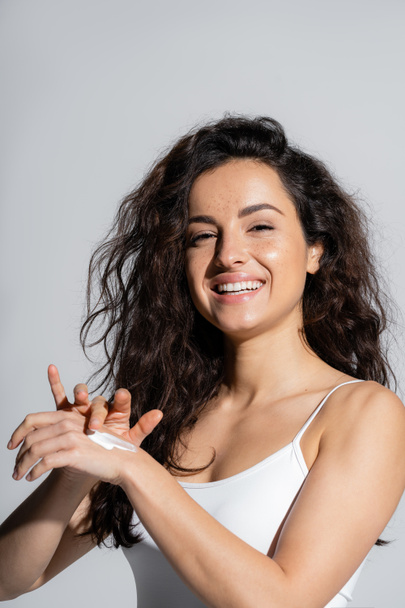 Cheerful woman in white top applying hand cream isolated on grey  - Foto, Imagem