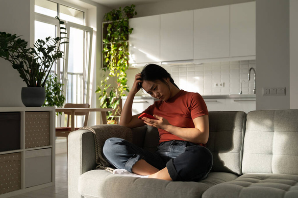 Depressed young Asian woman staring at mobile phone screen searching for reproductive health clinic online, sitting on sofa at home. Worried Korean girl reading information about medication abortion - Valokuva, kuva