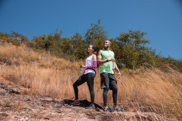 Athlete couple standing drink water while running in the forest, Refresh body by drinking water. African American man and woman runner sport training together. workouts and healthy lifestyle. - Фото, зображення