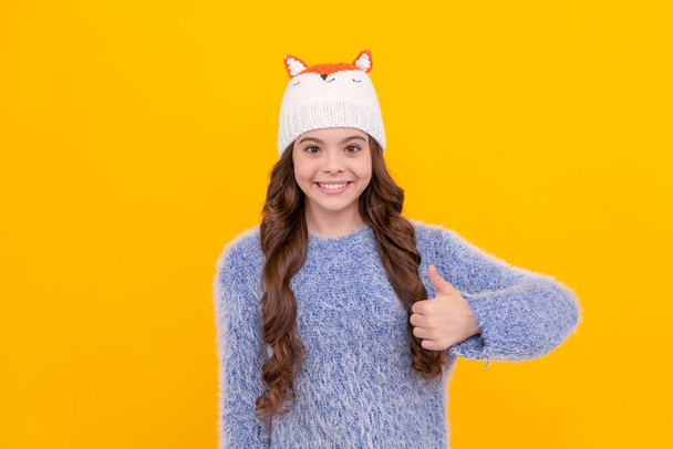 thumb up. positive kid with curly hair in hat. female fashion model. teen girl in knitwear on yellow background. portrait of child wearing warm clothes. express positive emotion. winter fashion. - Foto, Bild