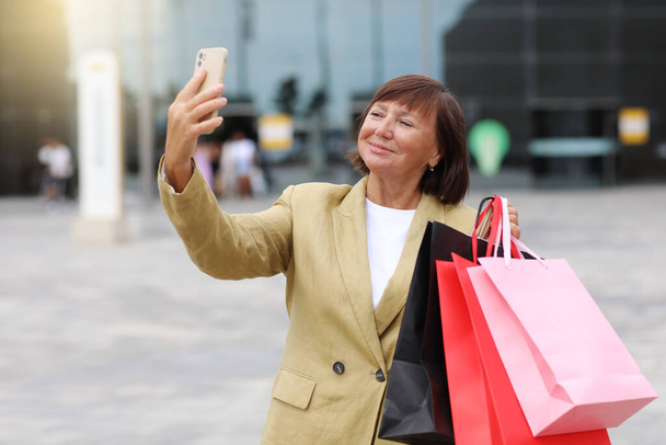 ショッピングバッグやスマートフォンを備えたスタイリッシュな大人の女性ブロガーは、ショッピングモールの近くのソーシャルネットワークでフォロワーと通信しています。消費主義、販売、豊かな生活、仮想生活の概念｜Black Friday. - 写真・画像