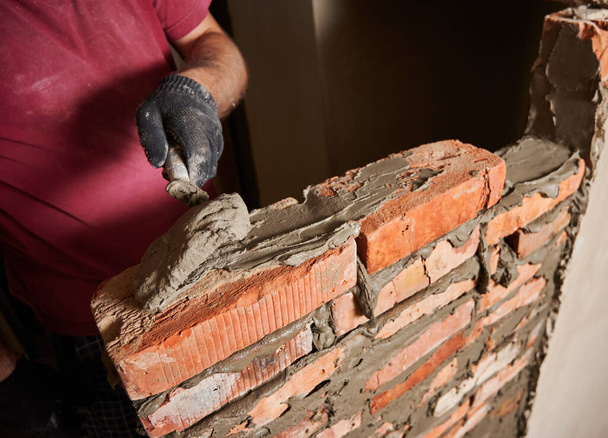 Close up of male hand in work glove laying brickwork in building under construction. Man bricklayer builder applying cement mortar on bricks with trowel tool. Masonry construction concept. - Φωτογραφία, εικόνα