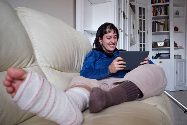 Young woman on couch at home with crutches and orthopedic watching series on tablet. Fracture of the broken leg foot or knee. Concept of rehabilitation and healing. Orthopedics and Traumatology. - Foto, imagen
