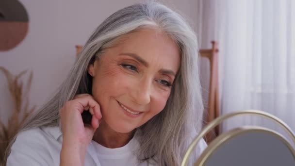 Anti-aging anti-wrinkled aging cosmetology. Old Caucasian woman senior mature aged lady female with perfect smooth complexion facial soft skin beautiful gray hair looking at mirror reflection smiling - Footage, Video