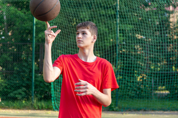 Cute young teenager in red t shirt with a ball plays basketball on court. Teenager dribbling the ball,  running in the stadium. Sports, hobby, active lifestyle for boys - Photo, Image