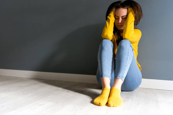 Full length of sad suffering young female in yellow sweater and socks touching head while sitting on floor near gray wall with closed eyes - Foto, Bild