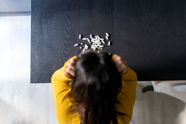 Top view of unrecognizable depress woman with long dark hair in yellow sweater touching head while sitting at table with pile of pills - Foto, Bild