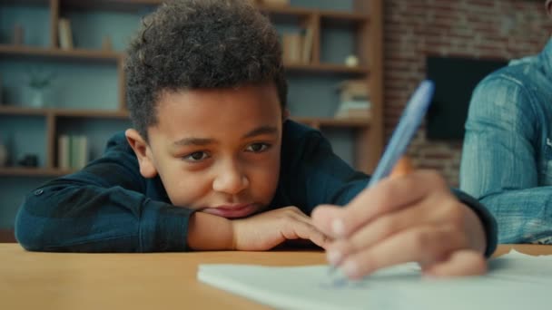 Tired exhausted sad upset fatigued little African American ethnic child boy kid son pupil schoolboy schoolchild lying on table desk writing homework boring class lesson drawing in notebook near father - Filmati, video
