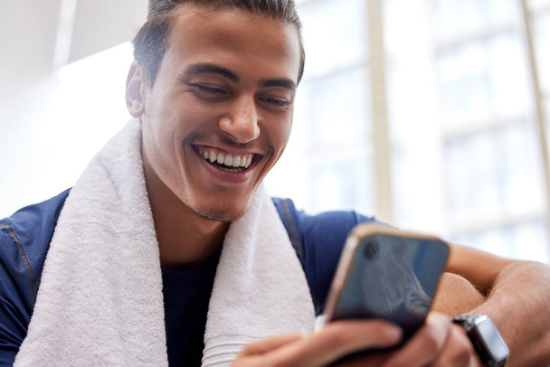 Man relax, fitness and smartphone in gym, smile and rest after workout, training and typing for social media. Male, athlete and guy with cellphone, break and connection for chatting and with towel. - Fotoğraf, Görsel