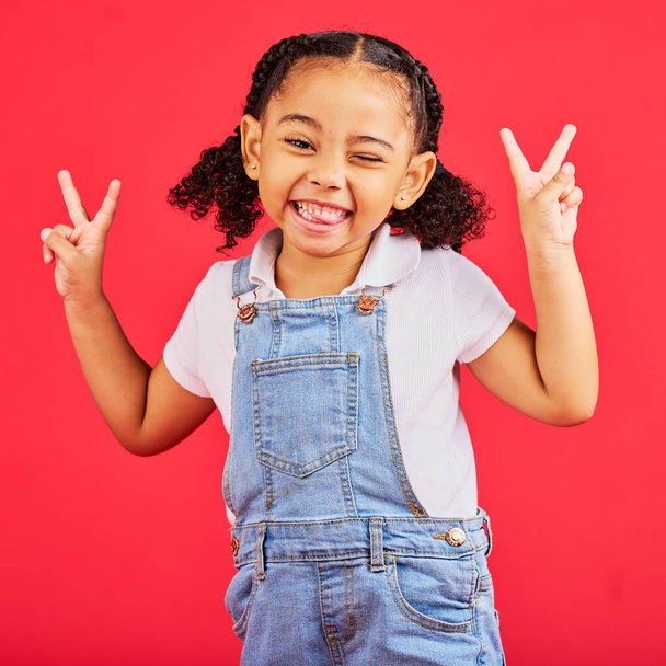 Happy, peace sign and wink with portrait of girl for summer, happiness and funny face. Meme, fashion and smile with child and hand sign for youth, comedy and positive in red background studio. - Valokuva, kuva