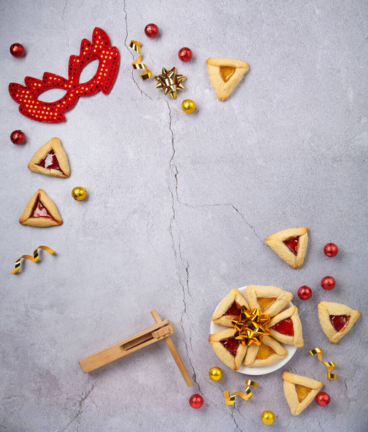 Purim celebration jewish carnival holiday concept. Tasty hamantaschen cookies, Triangular pastry, red carnival mask, noisemaker, sweet candies and party decor on gray stone table. Top view, flat lay, copy space. - Foto, afbeelding