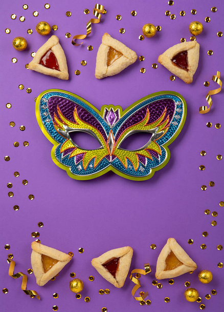 Purim celebration jewish carnival holiday concept. Tasty hamantaschen cookies, Triangular pastry, Festive carnival mask, noisemaker, sweet candies and party decor on purple Background. Top view, flat lay, copy space. - Photo, image