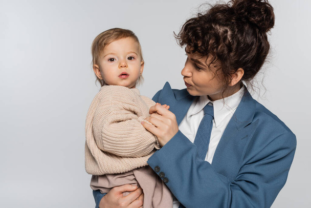 curly businesswoman in suit with tie looking at baby daughter isolated on grey  - Foto, Imagen