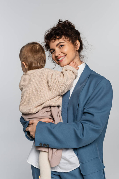 cheerful businesswoman in blue suit holding in arms baby girl isolated on grey  - Fotó, kép