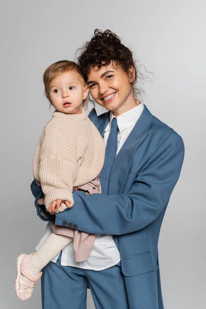 joyful businesswoman in blue suit holding in arms toddler daughter isolated on grey  - Photo, image