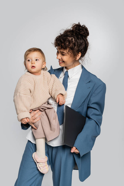 cheerful businesswoman in blue suit holding in arms toddler daughter and laptop isolated on grey  - Foto, imagen