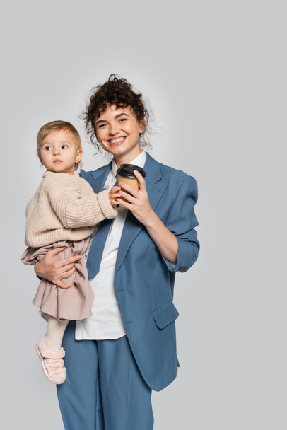 happy businesswoman in blue formal wear holding in arms toddler daughter and coffee to go isolated on grey  - Фото, изображение