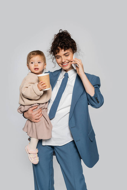 happy businesswoman in suit talking on smartphone while holding toddler daughter with takeaway drink isolated on grey  - Foto, immagini