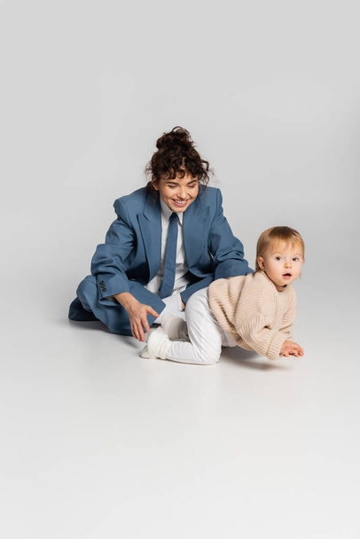 curly businesswoman in blue suit looking at toddler daughter crawling on grey background  - Zdjęcie, obraz