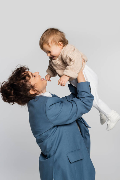 happy businesswoman in blue suit holding cheerful toddler girl in sweater isolated on grey  - Foto, afbeelding