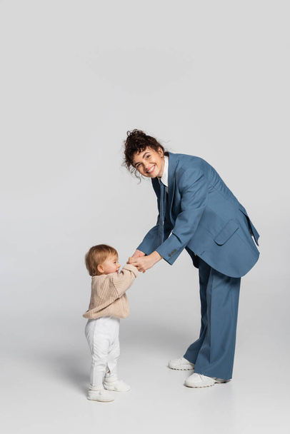 full length of happy businesswoman in blue suit holding hands with baby girl in sweater on grey  - Foto, Bild