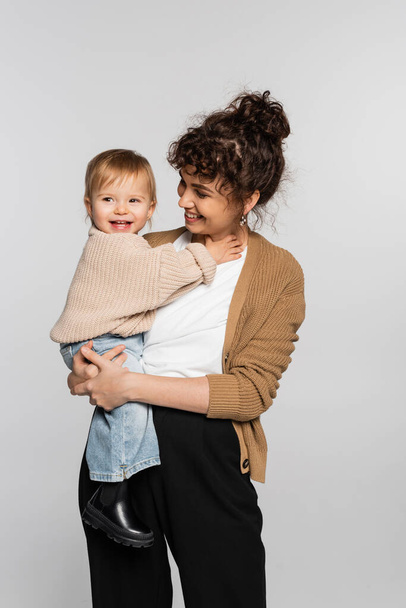 happy mother in casual clothes holding in arms cheerful baby girl in sweater isolated on grey  - Photo, Image