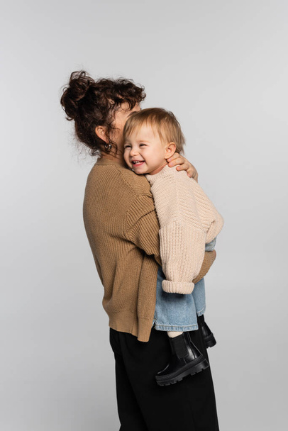 curly mother in casual clothes holding in arms happy baby daughter in sweater isolated on grey  - Foto, Imagen