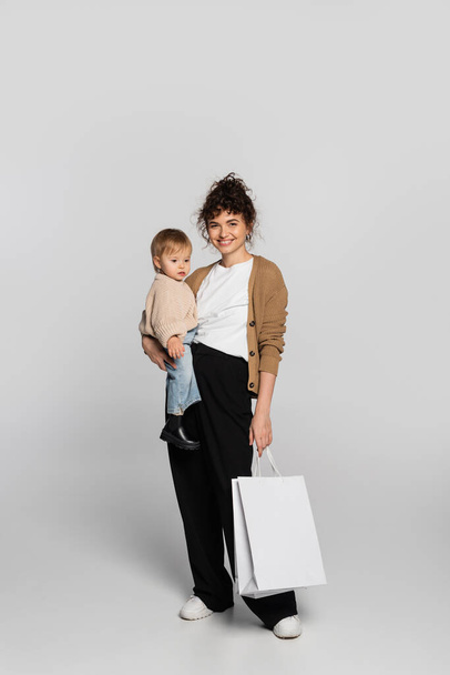 full length of happy mother in casual clothes holding in arms cheerful baby daughter and shopping bag on grey  - Foto, Imagem
