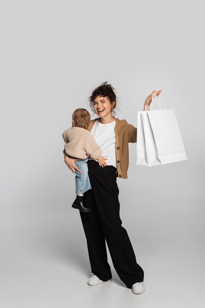 full length of cheerful mother in casual clothes holding in arms toddler baby and shopping bags on grey  - Valokuva, kuva