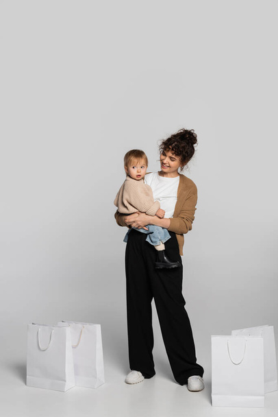full length of cheerful mother in casual clothes holding in arms toddler baby near shopping bags on grey  - Foto, Imagem