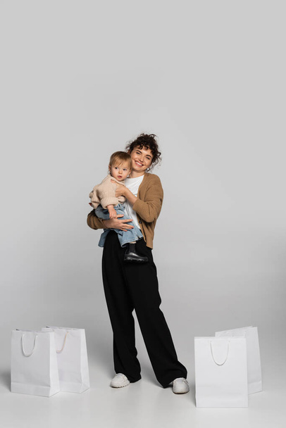 full length of cheerful mother in casual clothes holding in arms toddler girl near shopping bags on grey  - Фото, зображення