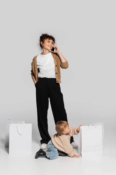 full length of happy woman in casual clothes talking on smartphone near toddler baby and shopping bags on grey  - Foto, immagini