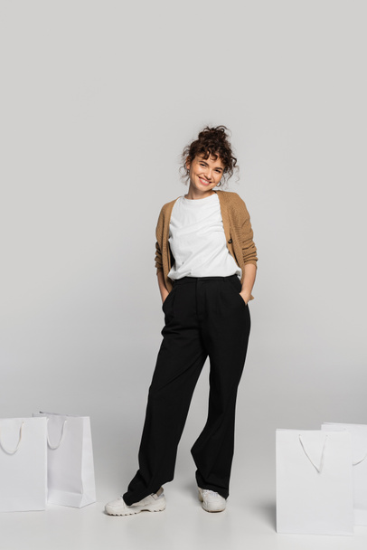 full length of cheerful woman in casual clothes standing with hands in pockets near shopping bags on grey  - Fotografie, Obrázek