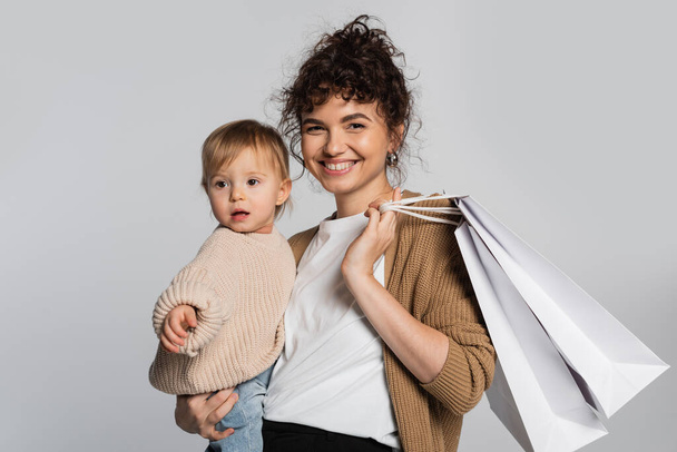 cheerful mother in casual clothes holding in arms toddler daughter and shopping bags isolated on grey  - Photo, Image