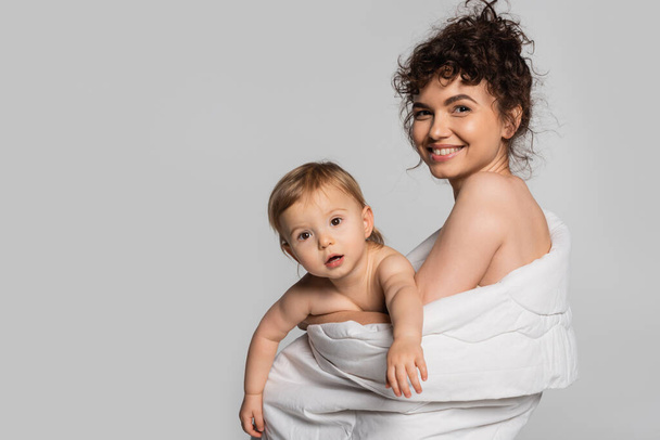 joyful mother and toddler baby girl covered in duvet isolated on grey  - Foto, afbeelding
