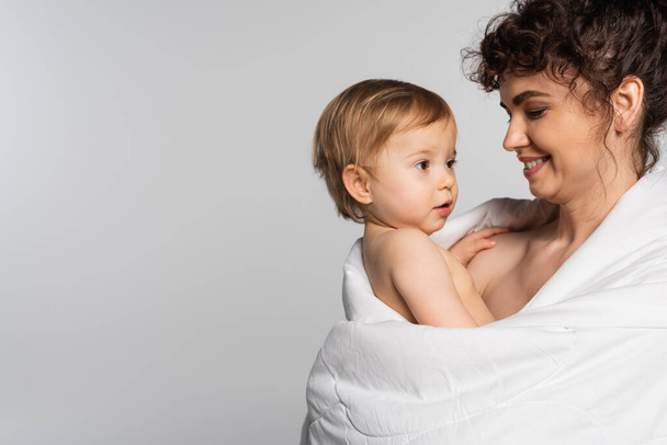 happy mother looking at toddler baby girl covered in duvet isolated on grey  - Foto, Imagem