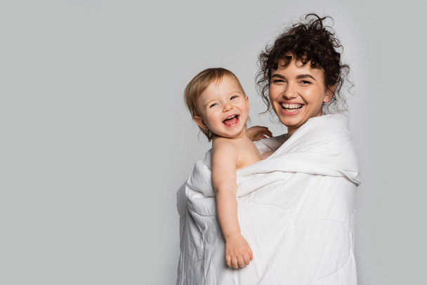 happy mother and positive toddler daughter covered in duvet isolated on grey  - Photo, Image