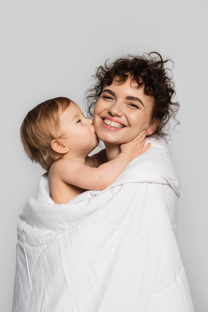 toddler baby girl kissing cheek of happy mother covered in white duvet isolated on grey  - Φωτογραφία, εικόνα