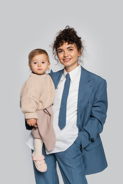 happy businesswoman in blue suit standing with hand in pocket and holding toddler daughter isolated on grey  - Zdjęcie, obraz