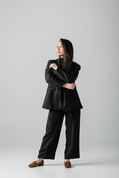 full length of young asian businesswoman in black suit standing and looking away on grey  - Foto, Imagem