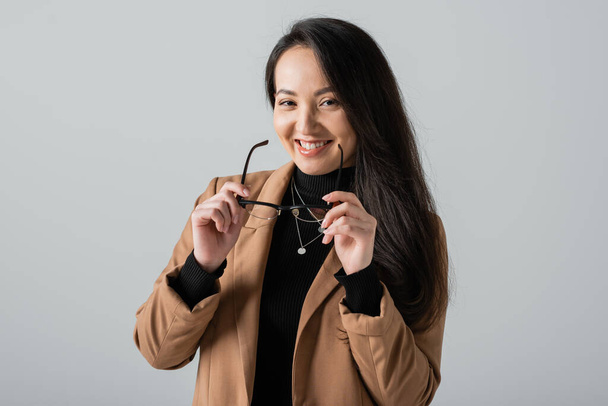 весела азіатська бізнес-леді в бежевому блістері тримає окуляри ізольовані на сірому
  - Фото, зображення