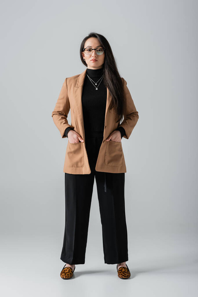 full length of young asian businesswoman in beige blazer and black pants standing with hands in pockets on grey  - Фото, зображення