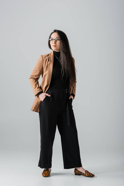 full length of confident asian businesswoman in beige blazer and black pants standing with hands in pockets on grey  - 写真・画像