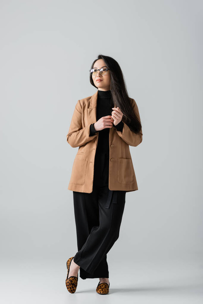 täyspitkä nuori aasialainen liikenainen beige bleiseri ja mustat housut seisoo ristissä jalat harmaa  - Valokuva, kuva