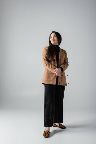 full length of brunette asian woman in beige blazer and black pants holding glasses on grey  - Photo, Image